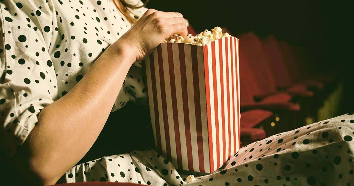 Decorative. A woman wearing a dress sits in a seat inside a darkened theater. She holds a large box of popcorn on her lap.
